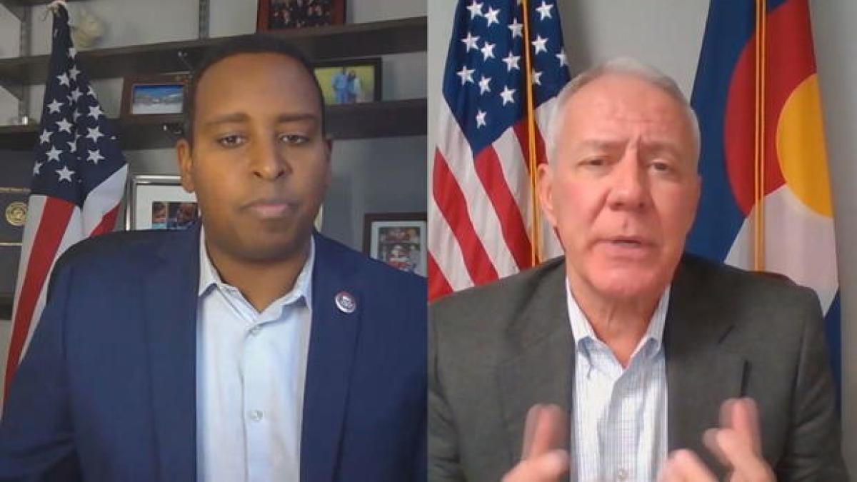 Representative Joe Neguse (left) and Representative Ken Buck (right) speak over a Zoom call. Both men are seated in their offices, where an American flag is visible in the background. 