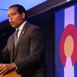 Congressman Joe Neguse stands in front of a podium while he gives a speech at the Colorado Democratic Party midterm watch party. A Colorado state flag is visible in the background. 